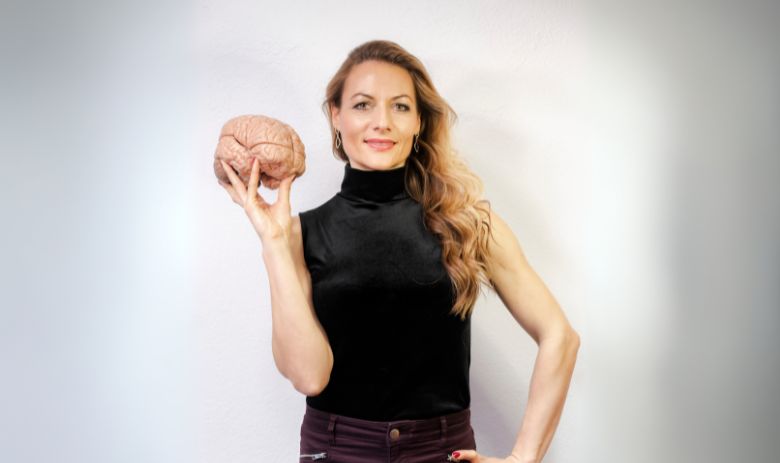 Barbara Studer Foto von Barbara Studer. Sie blickt mit einem selbstbewussten Lächeln in die Kamera. In der einen Hand hält sie das Modell eines Gehirns hoch, den anderen stützt sie an ihrer Hüfte.